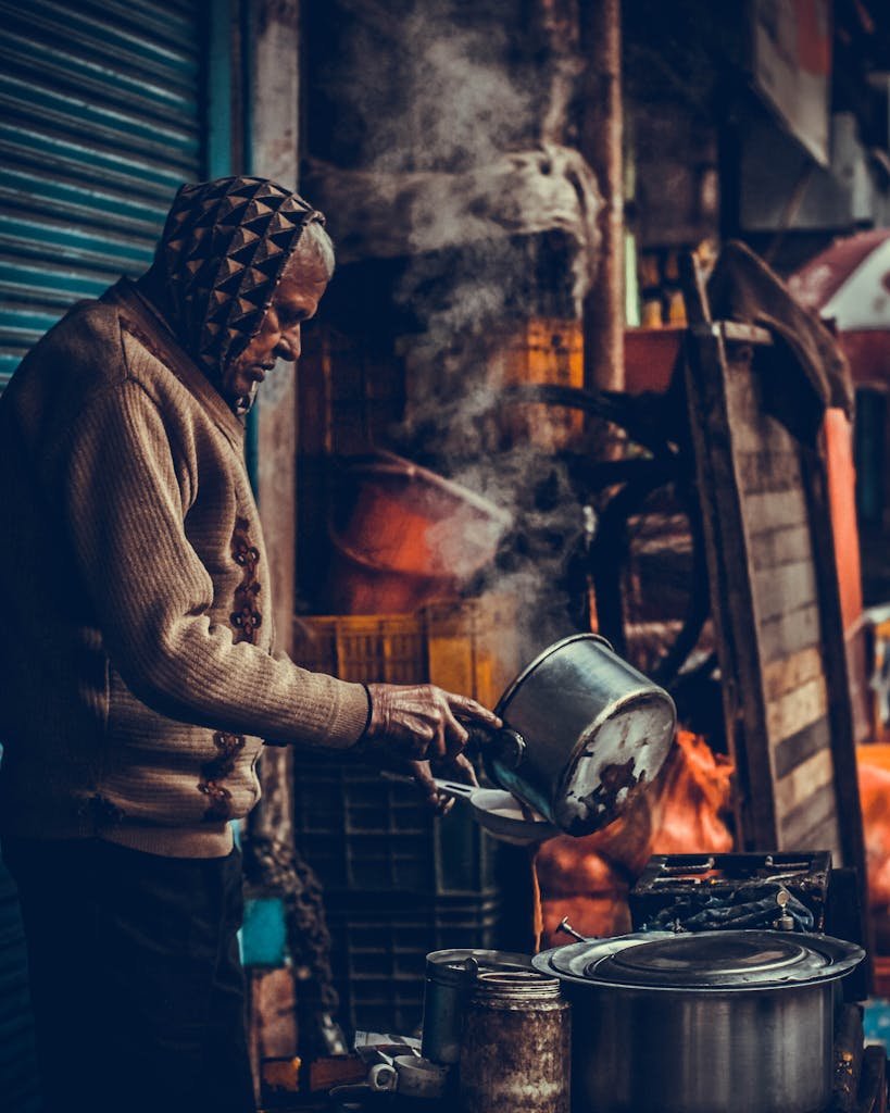 Senior man prepares food outdoors, steam rising from pot in bustling Srinagar street.